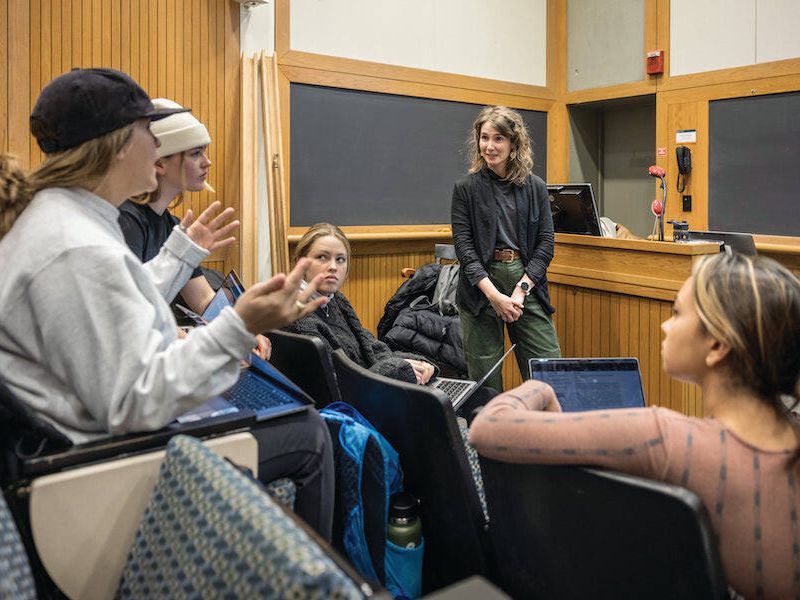 Instructor leading a discussion with students gathered around laptops in a wood-paneled Dartmouth lecture hall